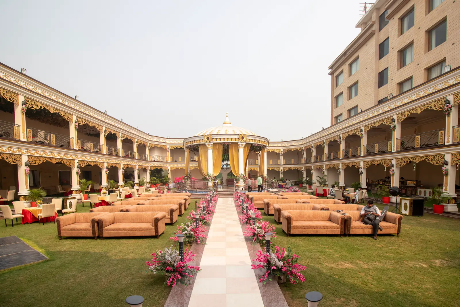 Lawn with sofa seating either side of a flower-lined aisle, surrounded by a two-storey colonnade